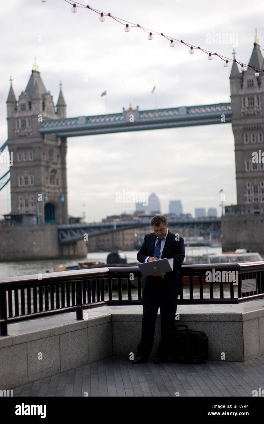 Uomo d'affari in piedi con un computer portatile nel Queens Walk accanto al Tamigi, con Tower Bridge sullo sfondo Foto Stock