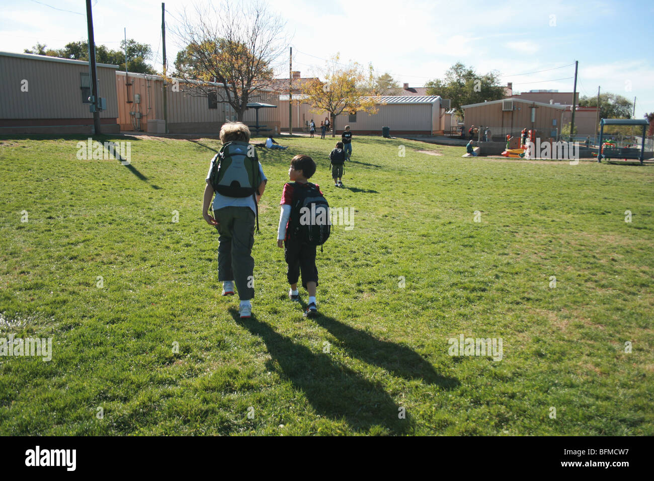 Due ragazzi camminare insieme a scuola sul campo erboso, scuola elementare, indossando zaini Foto Stock