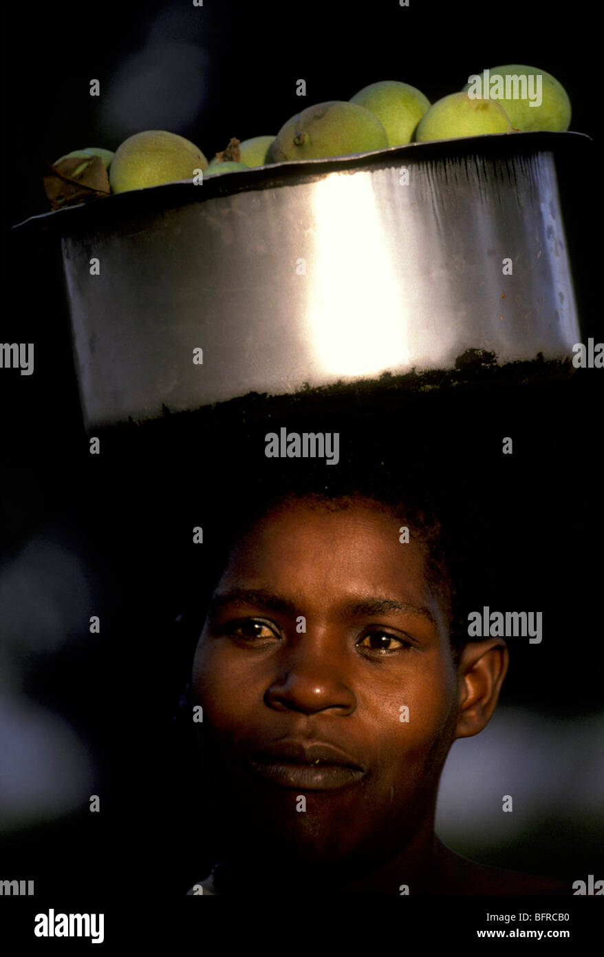 Ritratto di una donna Makua con green mango in una vasca in metallo bilanciato sulla sua testa Foto Stock