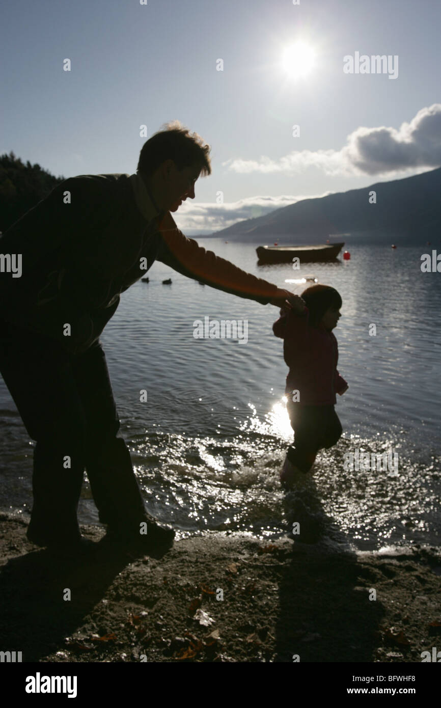 Area di Loch Lomond, Scozia. Vista profilarsi di una madre e figlia per mano mentre sguazzare nell'acqua. Foto Stock