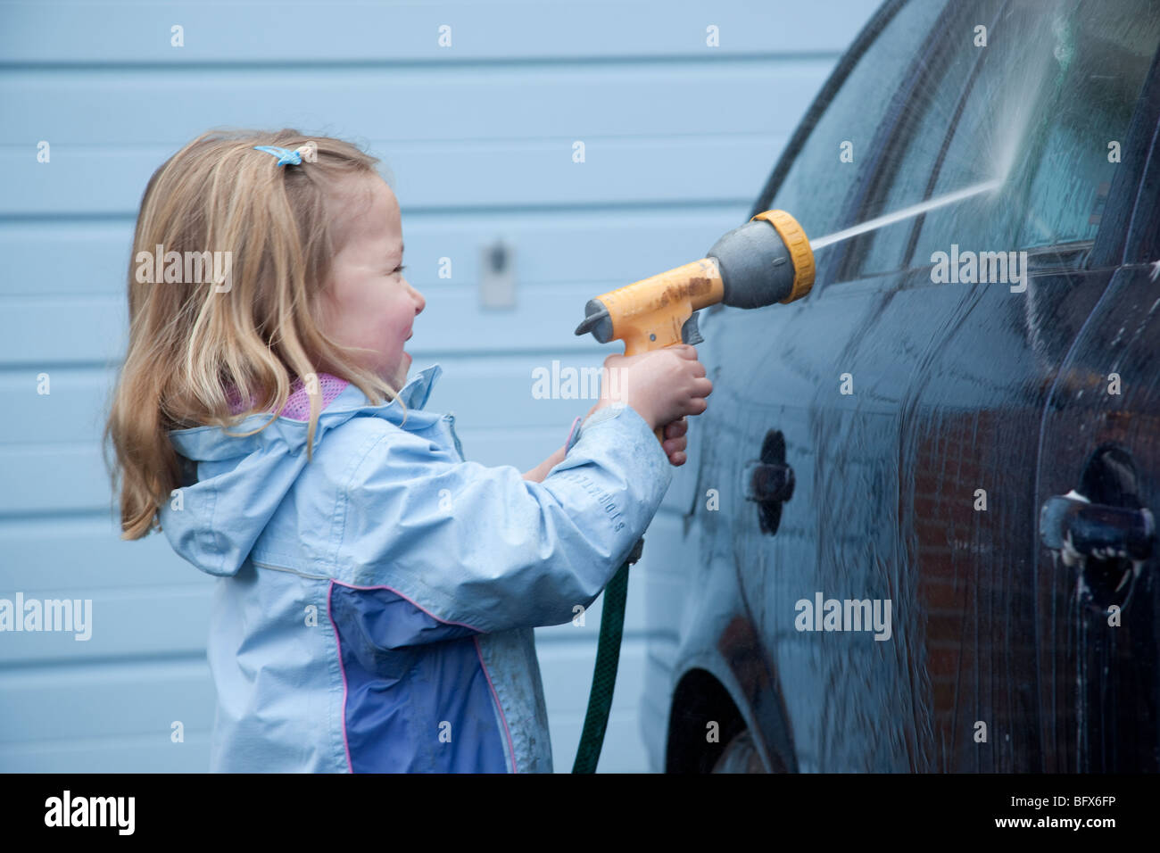 Macchina di lavaggio ragazza giovane figlia blu blu auto garage blue coat tubo flessibile rosa stivali da pioggia capelli biondi acqua carraio bambino Foto Stock