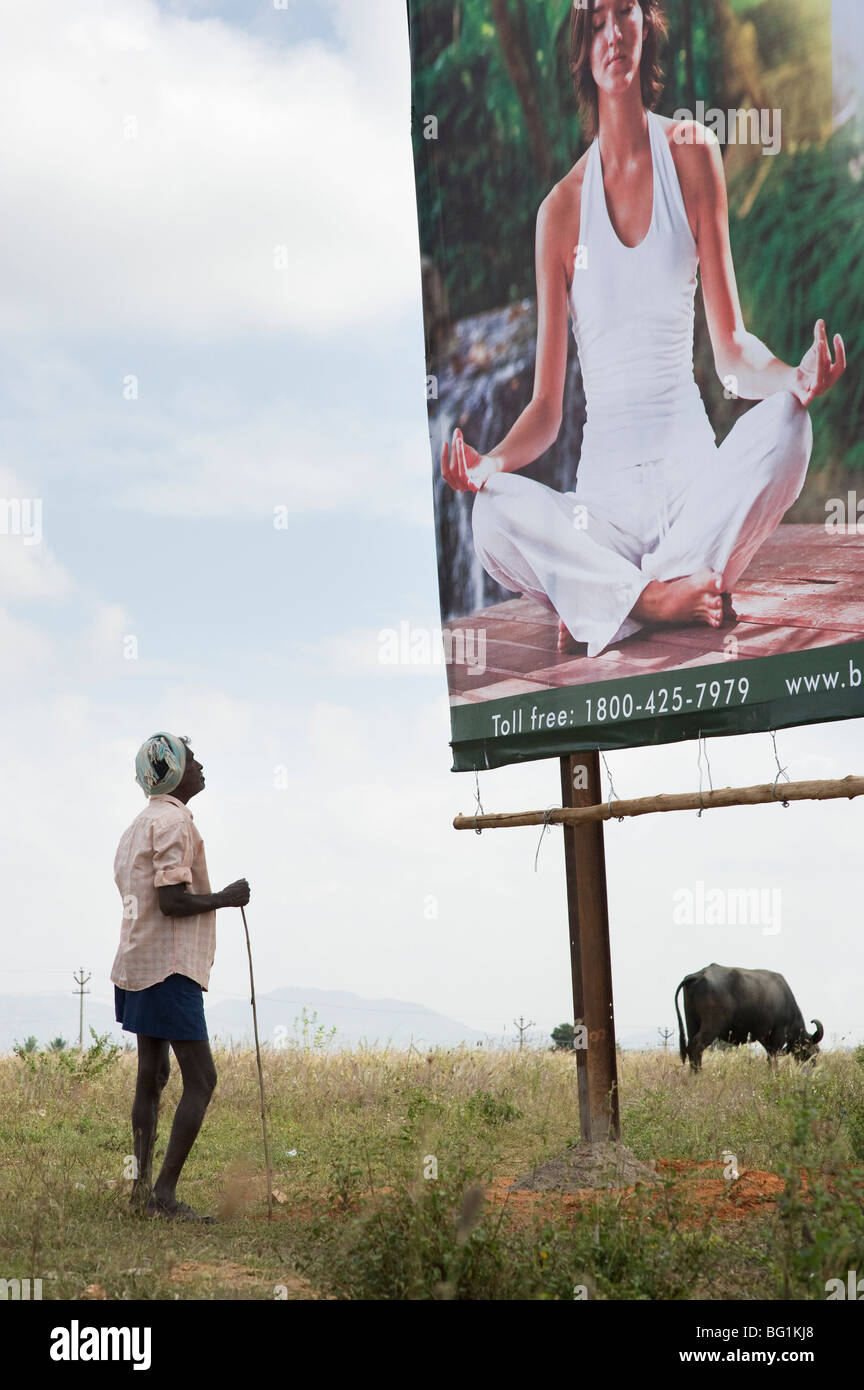 Il vecchio contadino indiano guardando uno stile occidentale Yoga meditazione billboard in campagna indiana. Andhra Pradesh, India Foto Stock