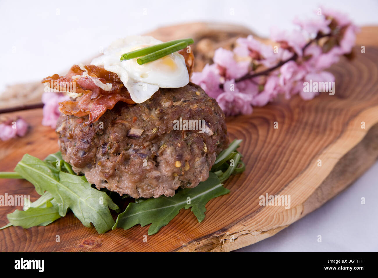 Burger gourmet su letto di rucola con pancetta e quaglie di uovo sulla parte superiore e servite sul piatto di legno con fiori selvatici. Foto Stock