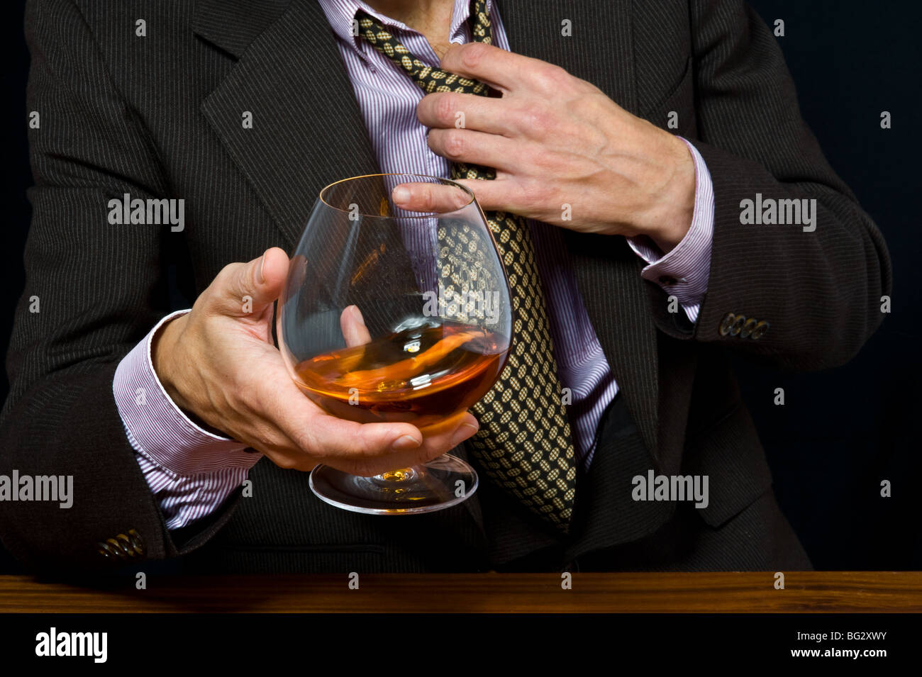 Vetro con alcool in una mano di un uomo in una tuta su sfondo scuro Foto Stock