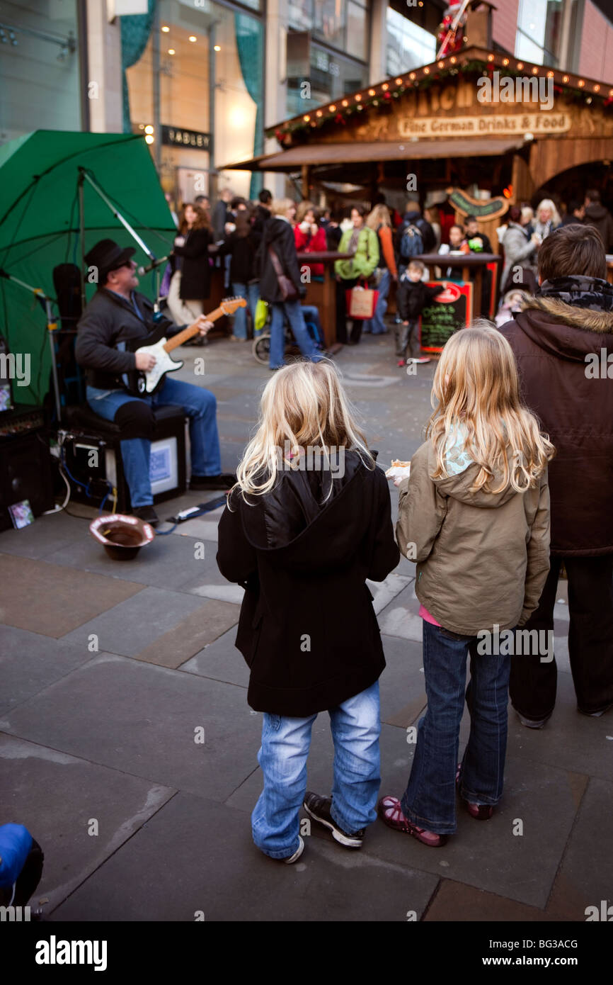 Regno Unito, Inghilterra, Manchester, Market Street, due giovani ragazze a guardare per suonare la chitarra blues busker Foto Stock