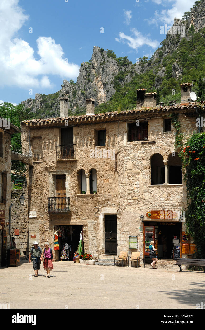 Coppia di turisti di mezza età camminano attraverso Piazza del Villaggio con negozi di souvenir, Saint Guilhem le Désert, Hérault, Languedoc Roussillon, Francia Foto Stock