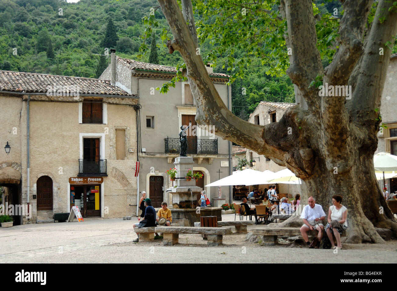 Coppia seduta sotto Plane Tree in Piazza del Villaggio, Place de la Liberté, Saint-Guilhem-le-Désert, Hérault, Languedoc Roussillon, Francia Foto Stock
