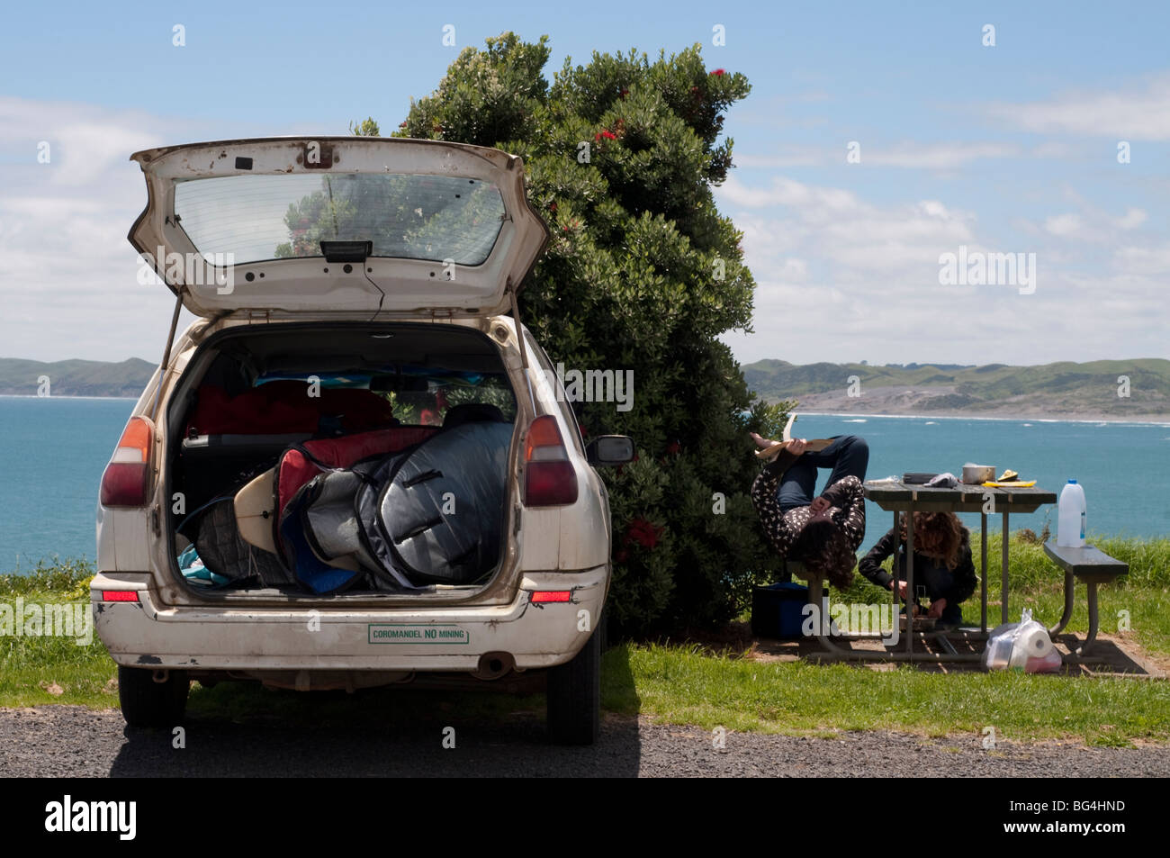 Stazione parcheggiata carro pieno di tavole da surf guardando oltre oceano, con due donne per la cottura e la lettura presi in Raglan, Nuova Zelanda Foto Stock