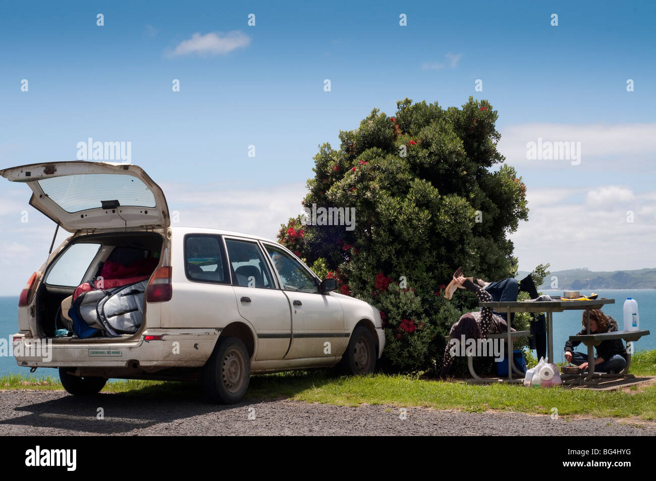 Stazione parcheggiata carro pieno di tavole da surf guardando oltre oceano, con due donne per la cottura e la lettura presi in Raglan, Nuova Zelanda Foto Stock