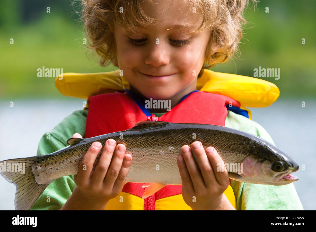 Un ragazzo giovane pescatore orgogliosamente esamina la trota arcobaleno ha pescato nel lago wit h la sua nuova canna da pesca su una sera d'estate. Foto Stock