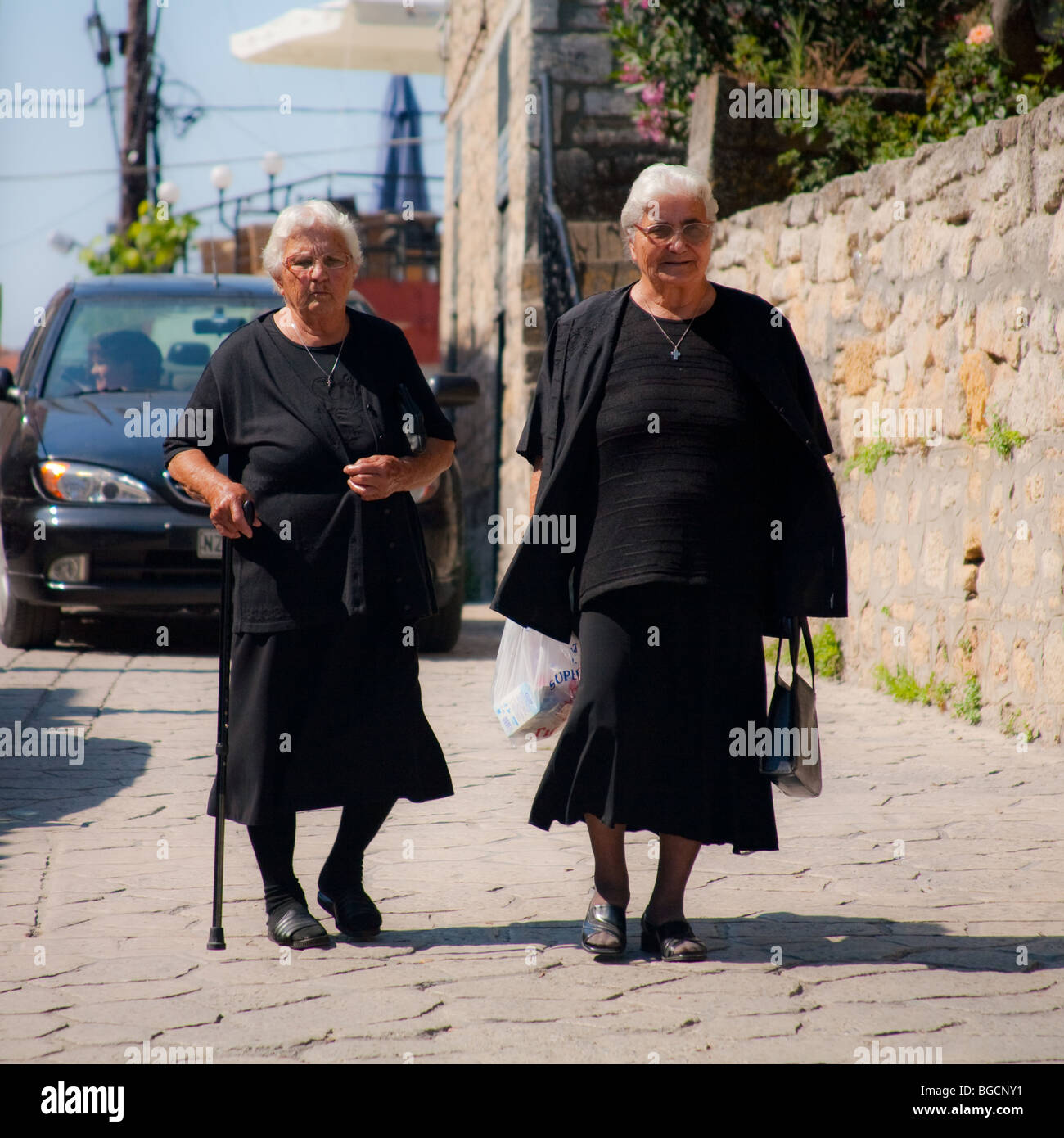 Due greci vedove camminare lungo una corsia vestita di nero BZH. Entrambi sono vecchi, uno ha un bastone da passeggio, sia trovare difficoltà a camminare. Foto Stock