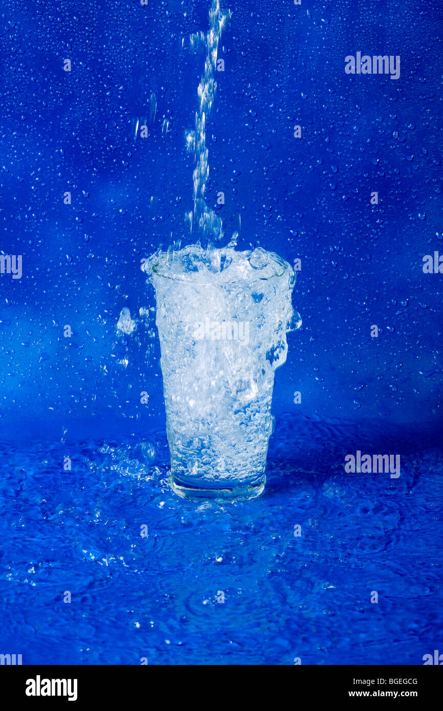 Versare acqua in un bicchiere e traboccante su uno sfondo blu Foto Stock