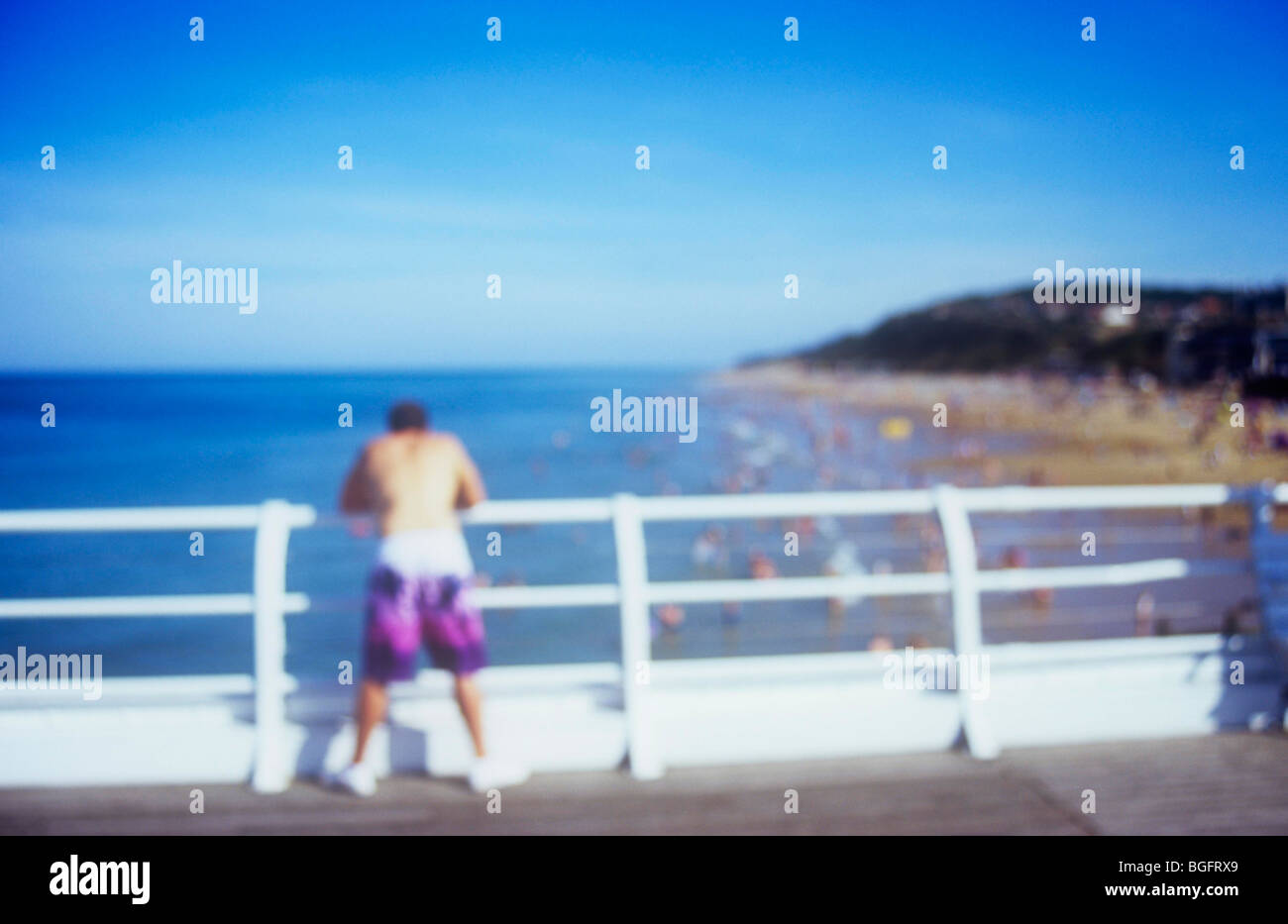 Vista impressionistica dell uomo appoggiato sulle ringhiere del molo e guardando fuori attraverso il mare e la spiaggia verso le scogliere Foto Stock
