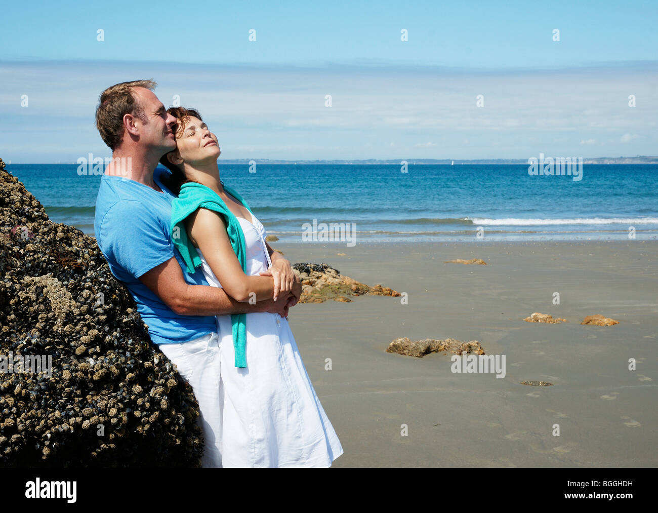 Giovane abbracciando sulla spiaggia, appoggiata contro una roccia, vista laterale Foto Stock