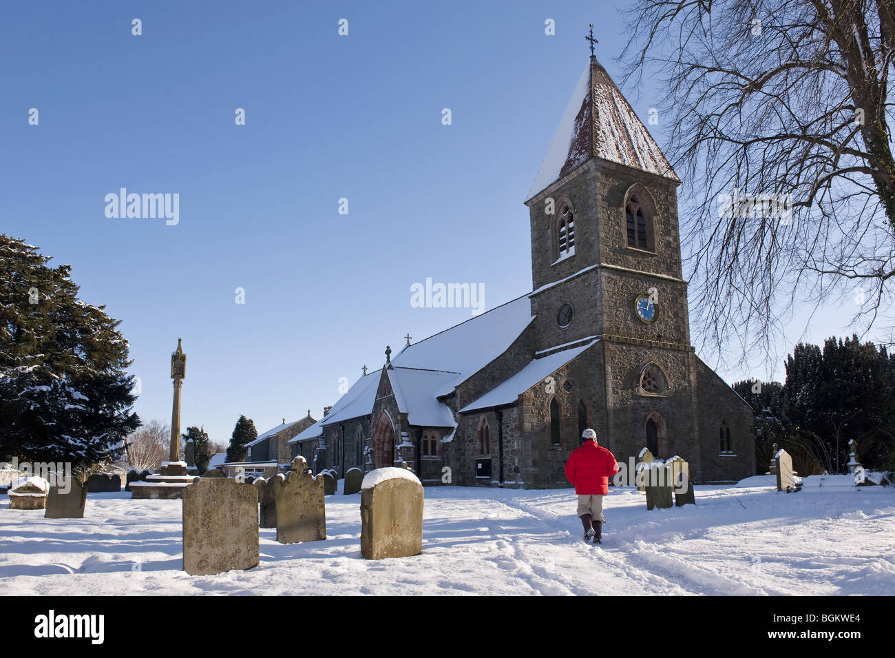 Persona solitaria cammina verso una coperta di neve pietra-walled village chiesa e cimitero commemorativo contro un cielo blu Foto Stock