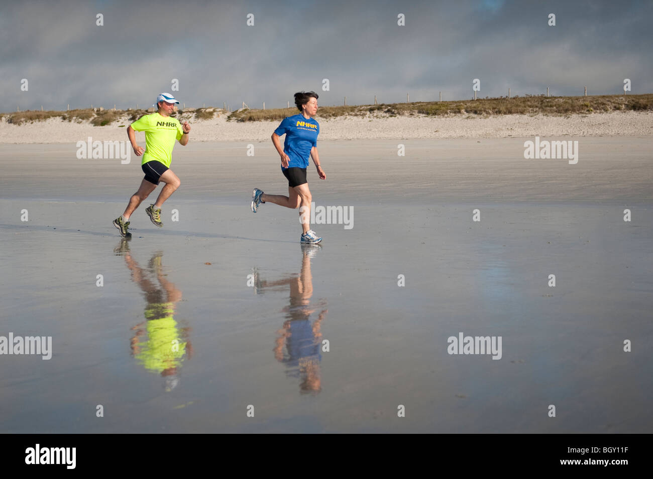 Due corridori prendere una corsa lungo una spiaggia di sabbia in Francia, loro vivacemente colorato t-shirts riflessa nella sabbia bagnata. su GR34 percorso Foto Stock