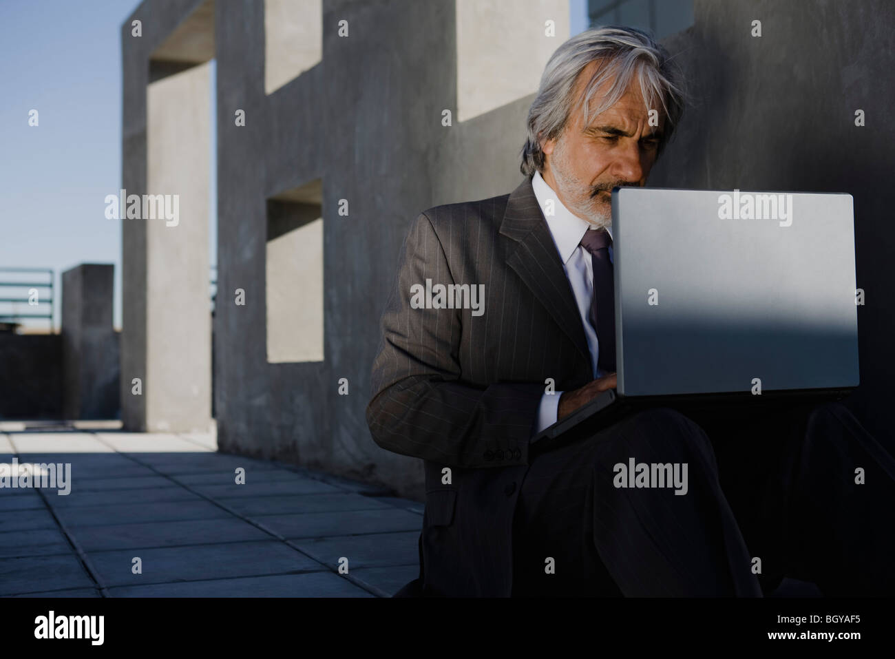 Uomo in tuta seduti sul marciapiede utilizzando laptop, edificio incompiuto in background Foto Stock