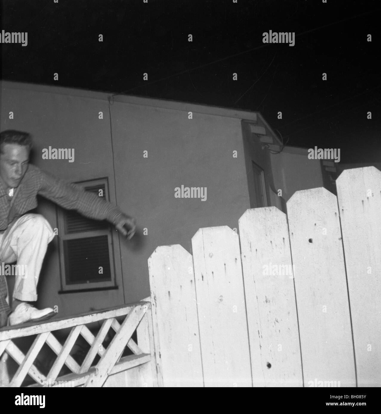 Una moda giovane uomo salta sopra un recinto accanto a una casa in una zona suburbana durante la fine degli anni cinquanta. Foto Stock