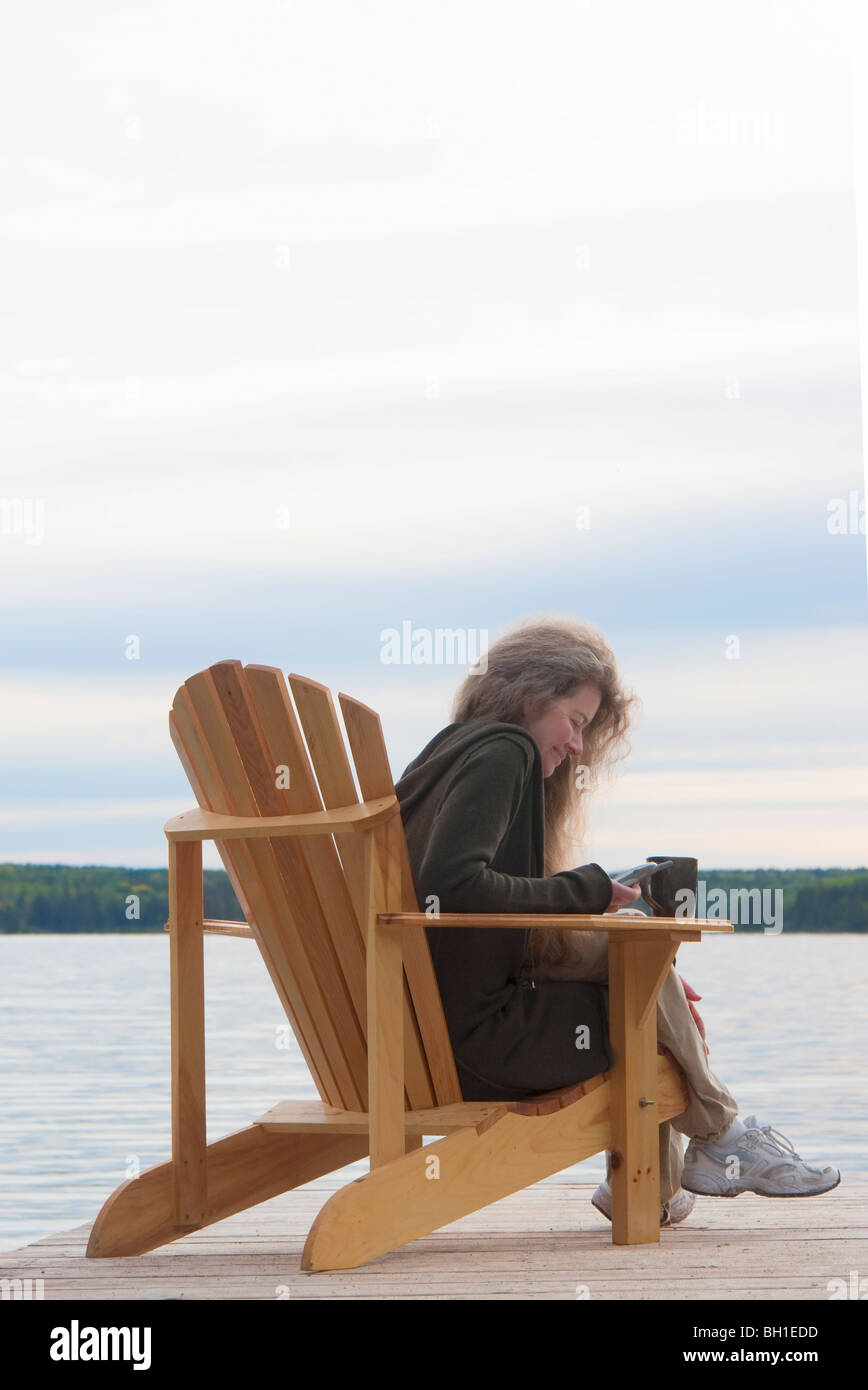 Donna di comporre sul cellulare, seduto nella sedia a sdraio sul dock, Clear Lake, Manitoba, Canada Foto Stock
