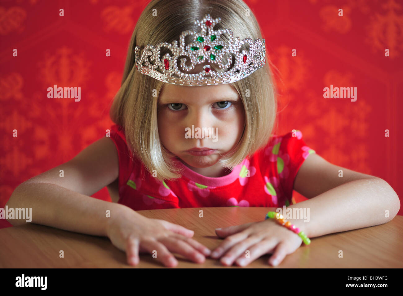 Giovane ragazza che indossa un abito rosso e un Princess Crown seduti ad un tavolo, accigliata con una misera espressione Foto Stock