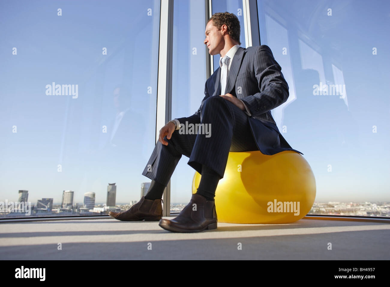 Un imprenditore seduto su una sfera svizzero nell'angolo di un ufficio guardando fuori della finestra attraverso i grattacieli Foto Stock