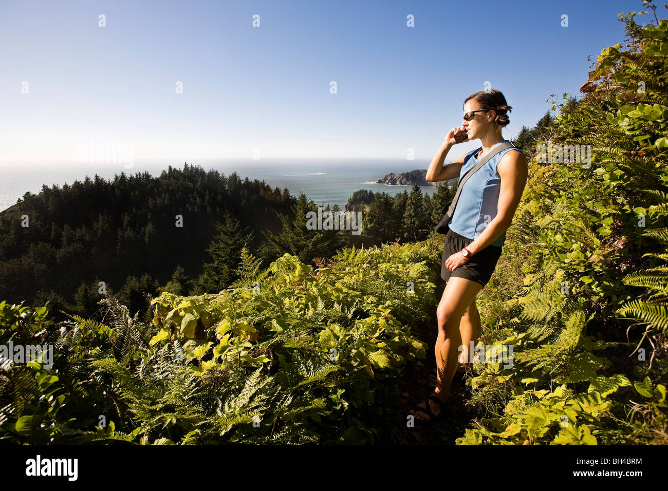 Una donna con occhiali da sole e vestiti estivi escursioni un sentiero costiero in Northwest Oregon e pause per rispondere il suo telefono cellulare. Foto Stock