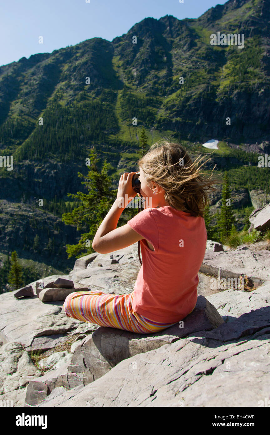 Una giovane ragazza sorge su un promontorio roccioso e si guarda attraverso il suo binocolo nel Parco Nazionale di Glacier, Montana. Foto Stock