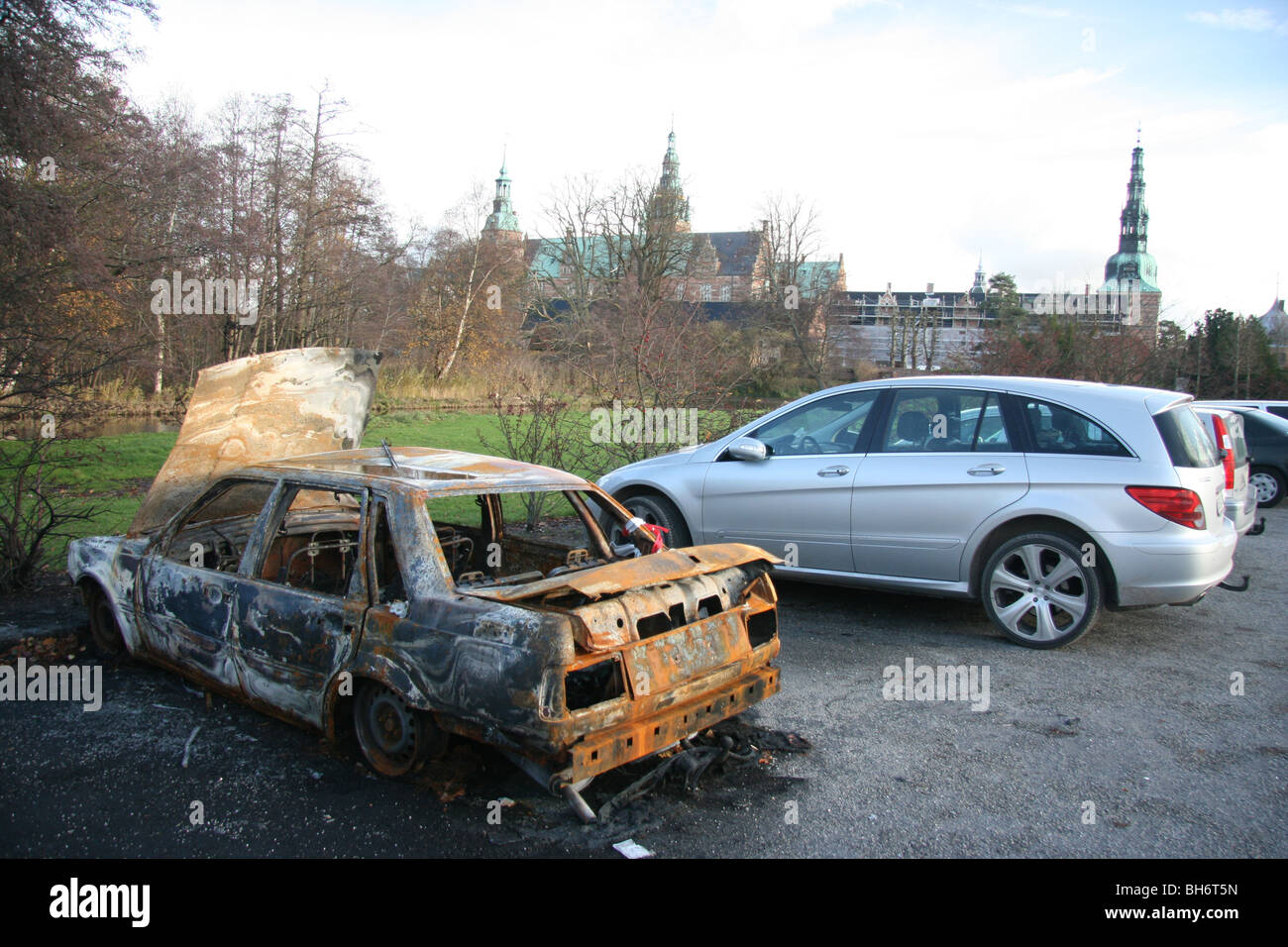 Un bruciato auto in contrasto con il nuovo modello di auto e il castello nel parcheggio del Castello Frederiksborg, Hilleroed, Danimarca. Foto Stock