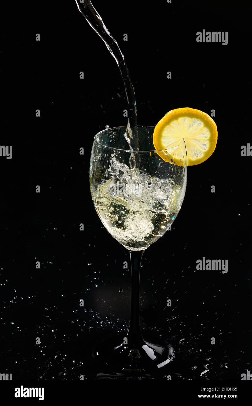 Versare il vino bianco schizzi in un bicchiere con una fetta di limone su sfondo nero Foto Stock