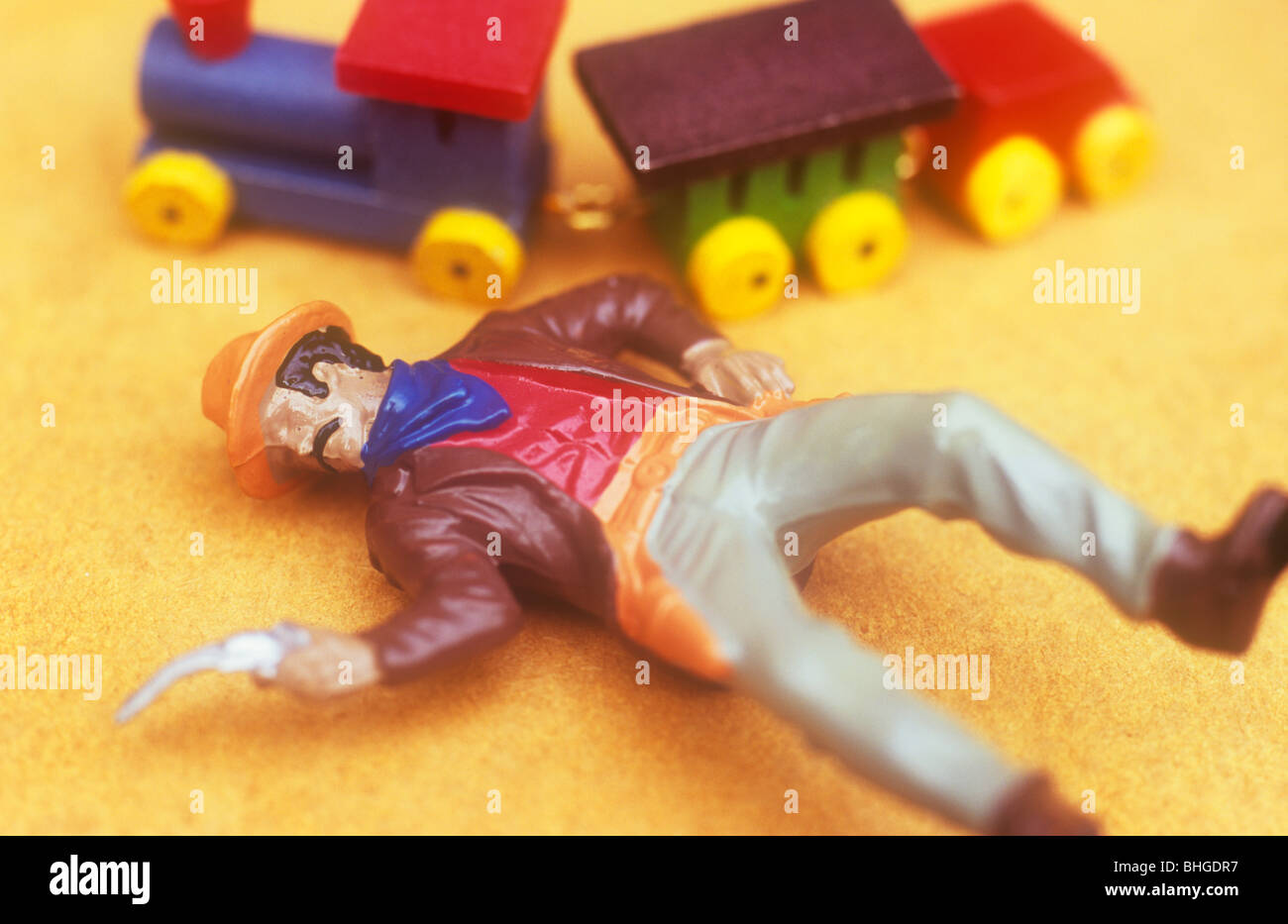Chiusura del modello di cowboy pistola di puntamento e stare sdraiato sulla schiena su sfondo beige con piccoli treni di legno e carrelli dietro Foto Stock
