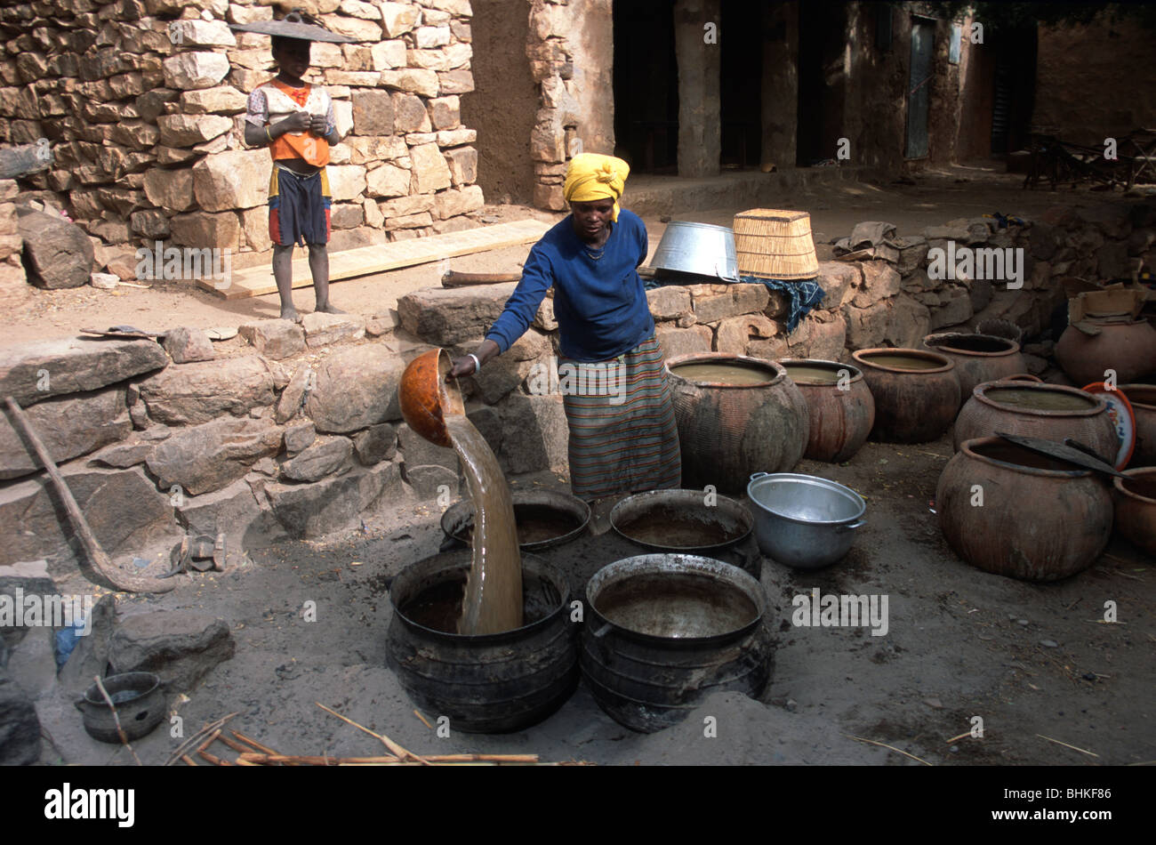 Una donna del Mali versando in parte di infuso di birra di miglio in una pentola, in Tereli, Mali, mentre un ragazzo di orologi con un coperchio sulla sua testa. Foto Stock