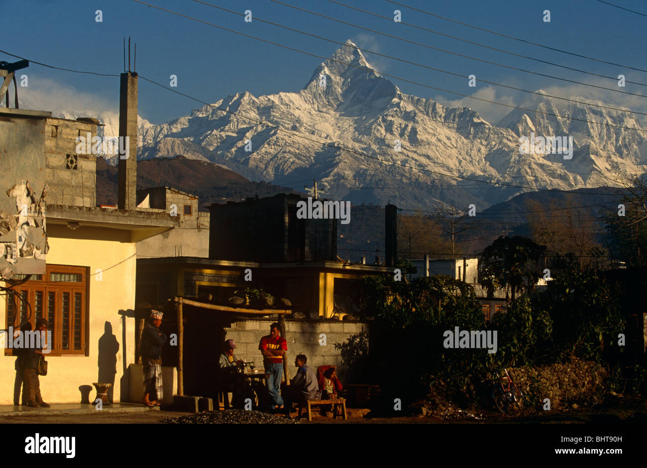 Al di sotto della snow-capped picco di Machapuchare montagna, un gruppo di gente nepalese chat su bassa sedi al di fuori della loro casa. Foto Stock