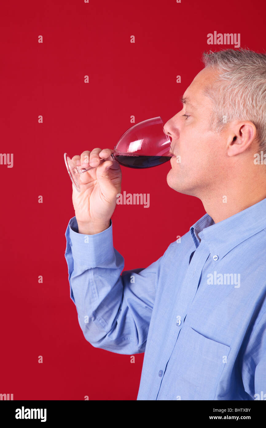 Uomo in una camicia blu di bere un bicchiere di vino rosso contro uno sfondo di colore rosso. Foto Stock