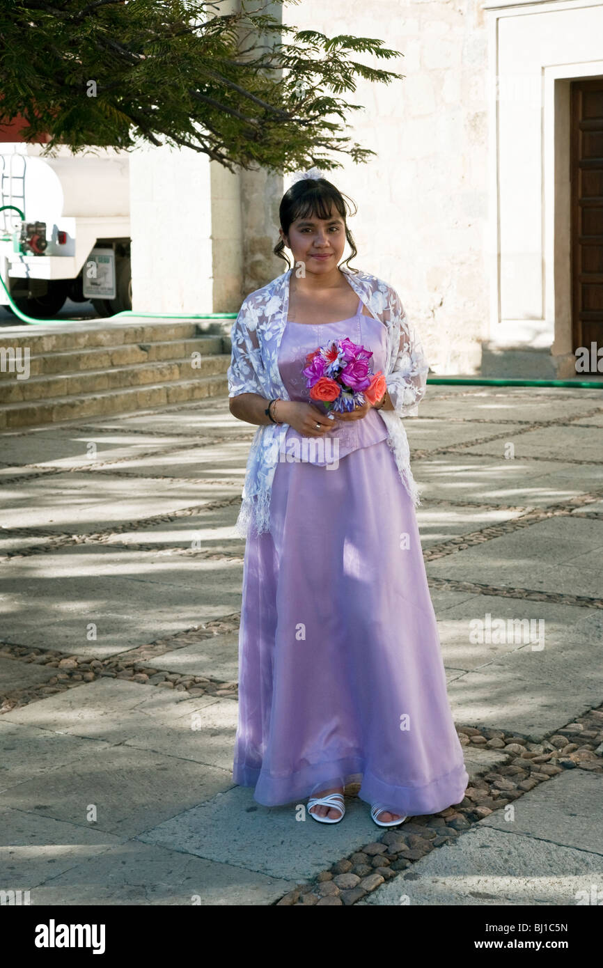 Pretty teen messicano in piedi in plaza al di fuori di chiesa di Santo Domingo che indossa camice per la sua cerimonia Quinceanera & holding fiori Foto Stock