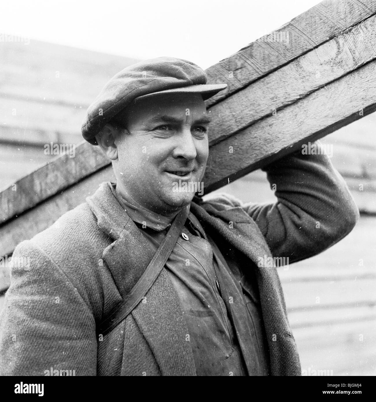 Inghilterra, 1950s. Close-up di un lavoratore per il London Brick Company, che indossa una giacca e cappuccio, portante tre tavole di legno. Foto Stock