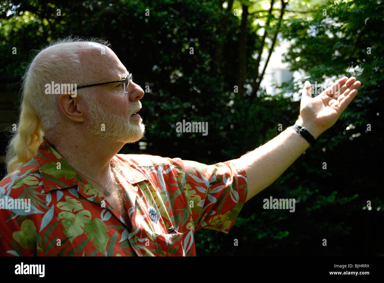 Capelli bianchi gentleman con la barba e coda di cavallo che indossa camicia hawaiana si estende il braccio o le onde nel gesto di amicizia in giardino Foto Stock