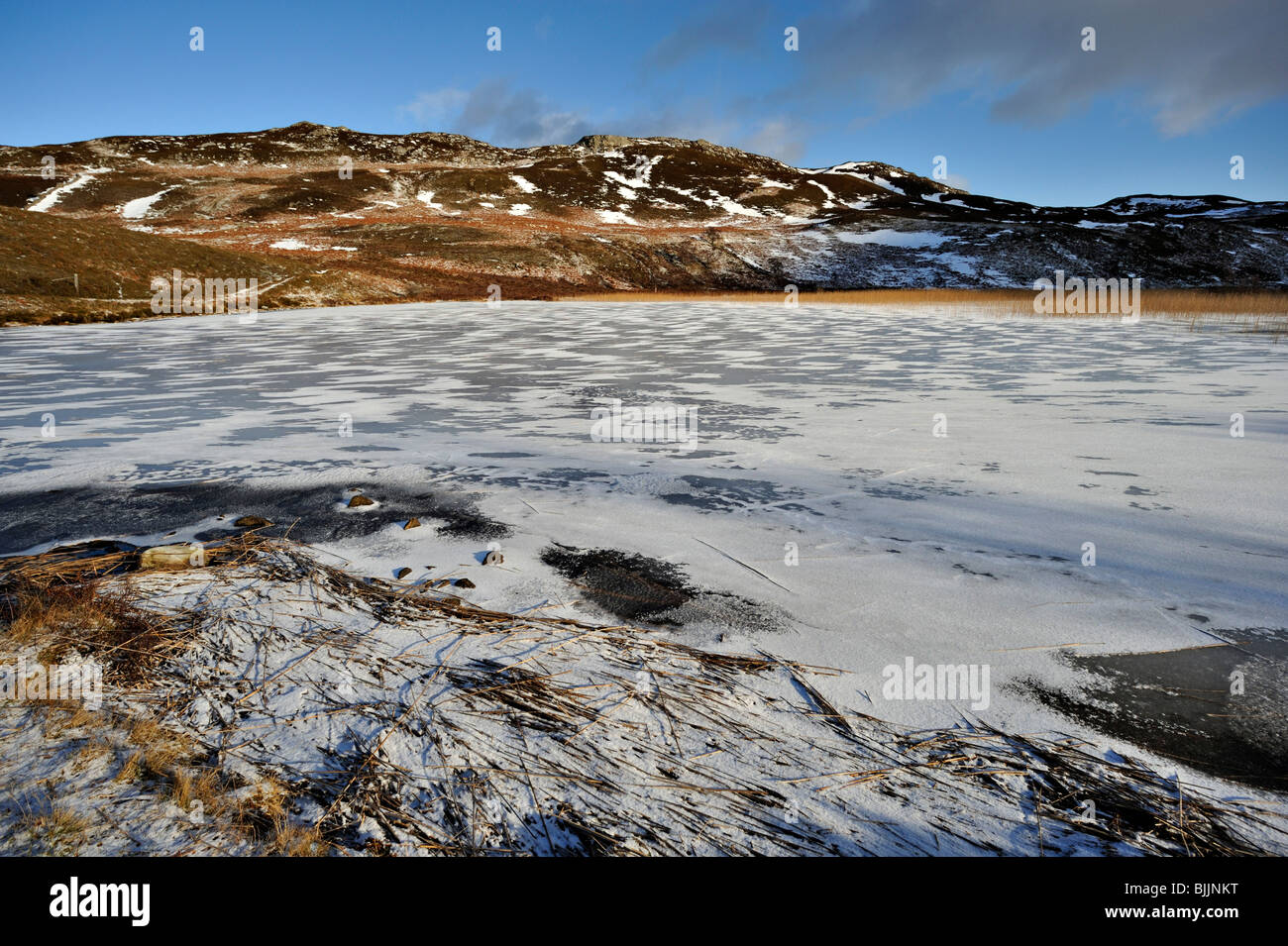 Soleggiato lago ghiacciato di ghiaccio, neve, canne e basse colline con frammentaria della neve Foto Stock
