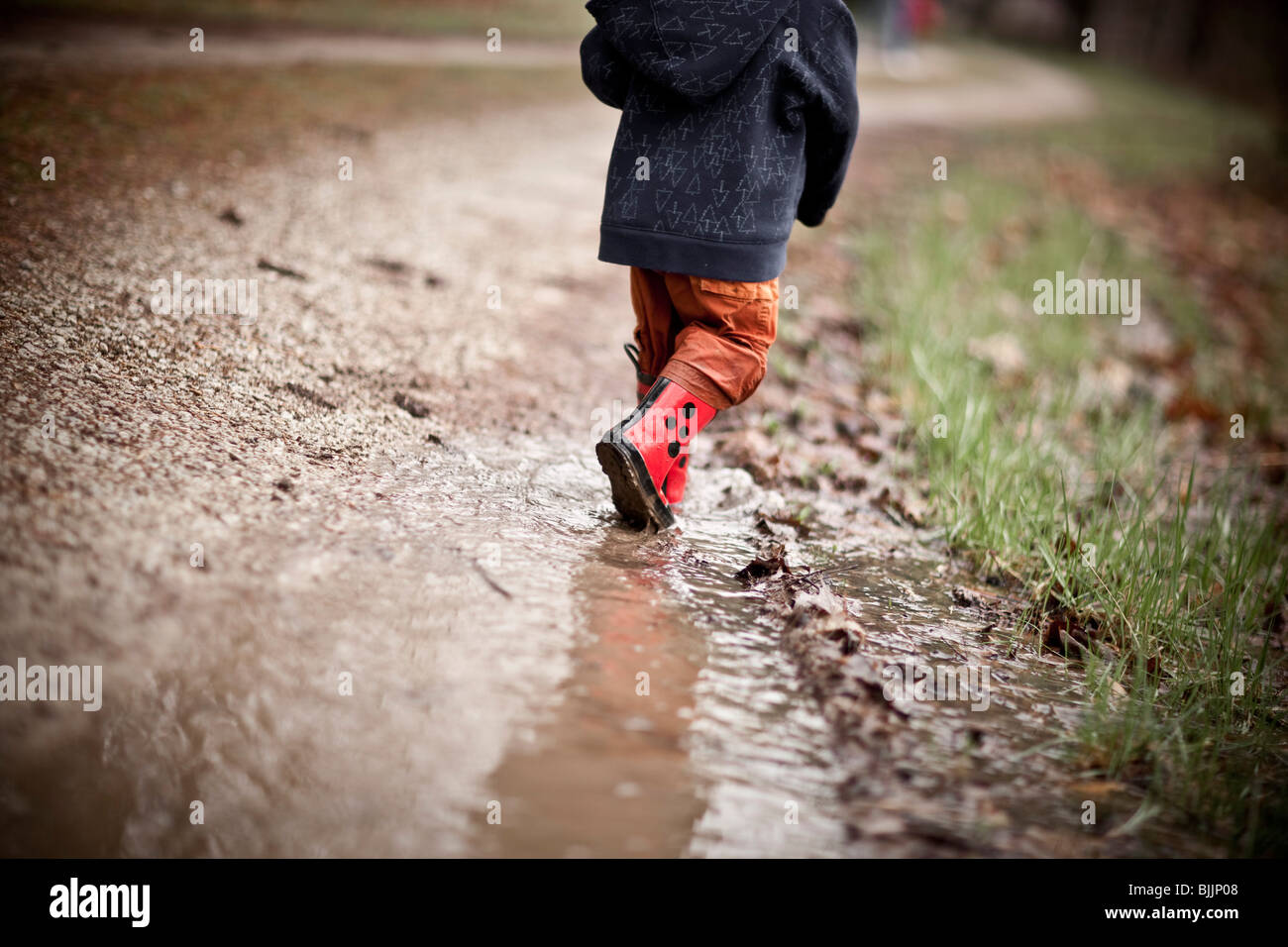 Giovane ragazzo indossare stivali da pioggia passeggiate attraverso una pozza d'acqua. Foto Stock