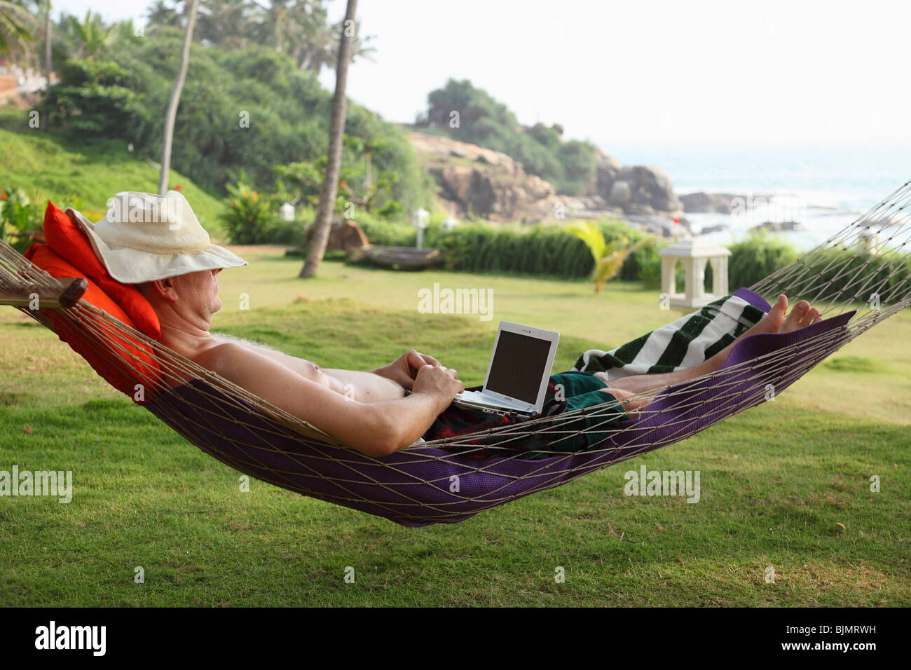 Uomo che indossa un cappello, giacente in una amaca e rilassarsi mentre si lavora con un netbook, Betsaida eremo vicino a Kovalam, Kerala, sout Foto Stock