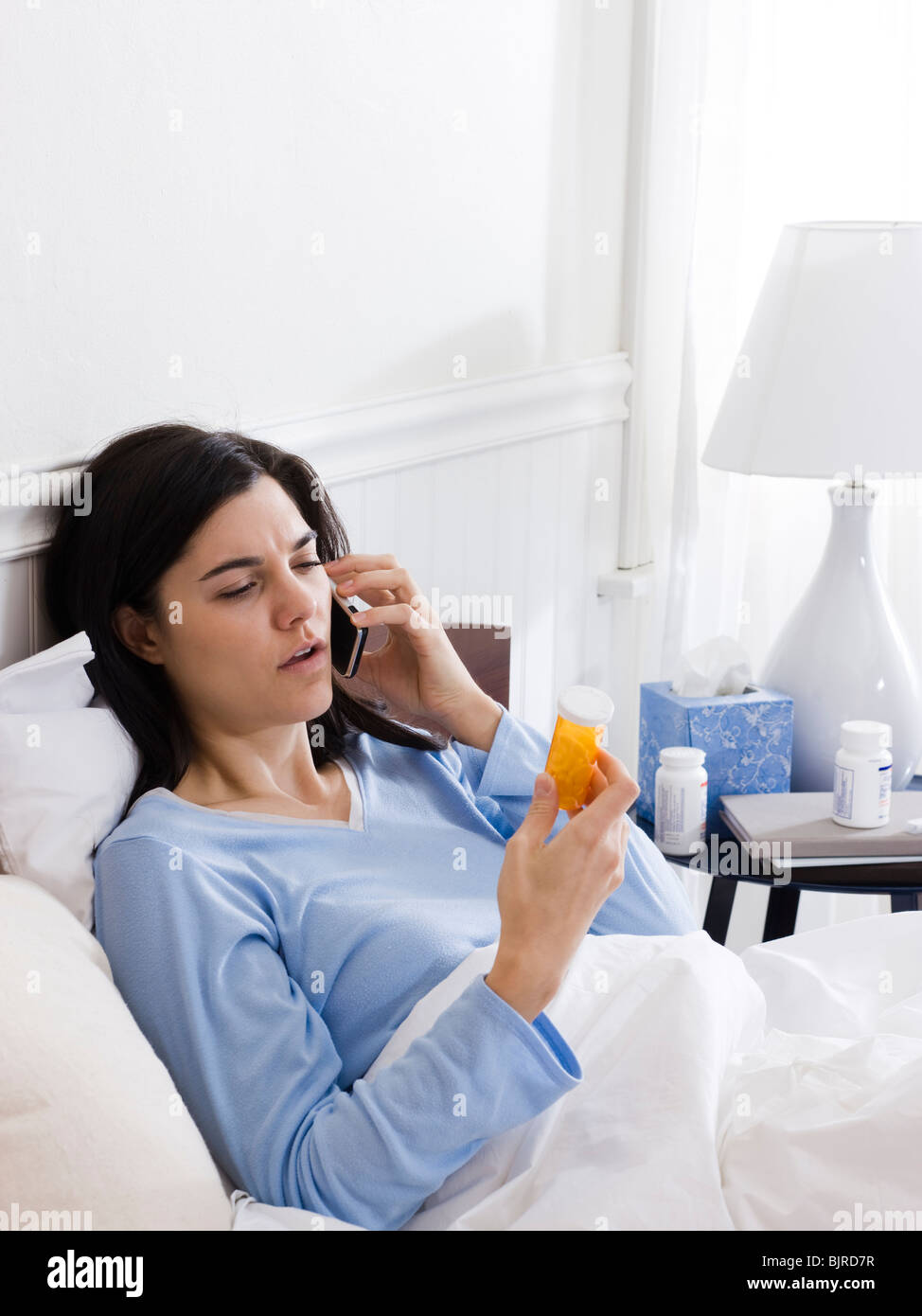 Orem, Utah, Stati Uniti d'America, giovane donna malata nel letto guardando pillola bottiglia e parlando al telefono Foto Stock