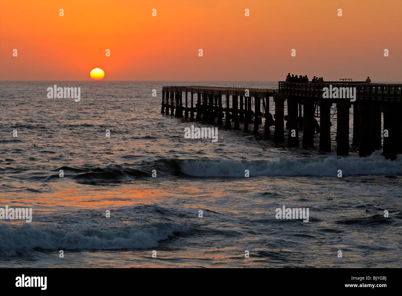 Vista di una profila molo costiere con persone a guardare un tramonto incandescente Foto Stock