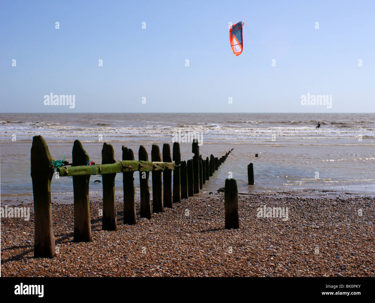 Kite Surfer usando un vivacemente colorato aquilone al potere lui lungo. La verticale di legno posti in primo piano sono vecchi e le difese del mare. Foto Stock