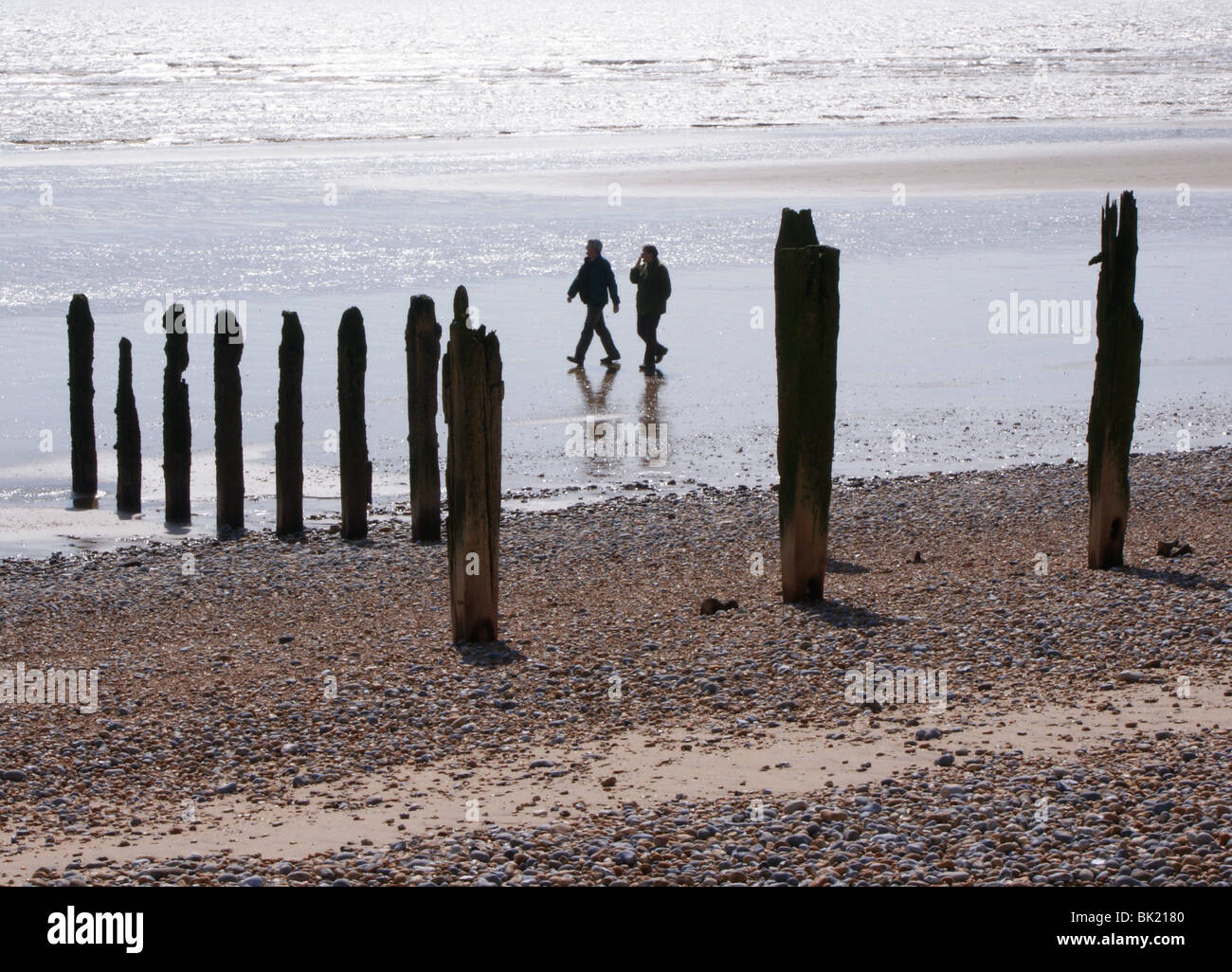 Due persone in la distanza a piedi su una spiaggia in East Sussex. Il montante posti di legno sono i resti della vecchia le difese del mare. Foto Stock