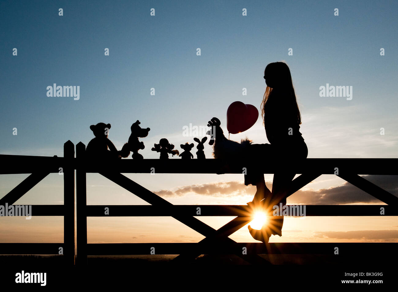 Ragazza con un cuore palloncino con una rag doll, pollo, la volpe, il coniglio e orso peluche seduto su un cancello al tramonto. Silhouette Foto Stock