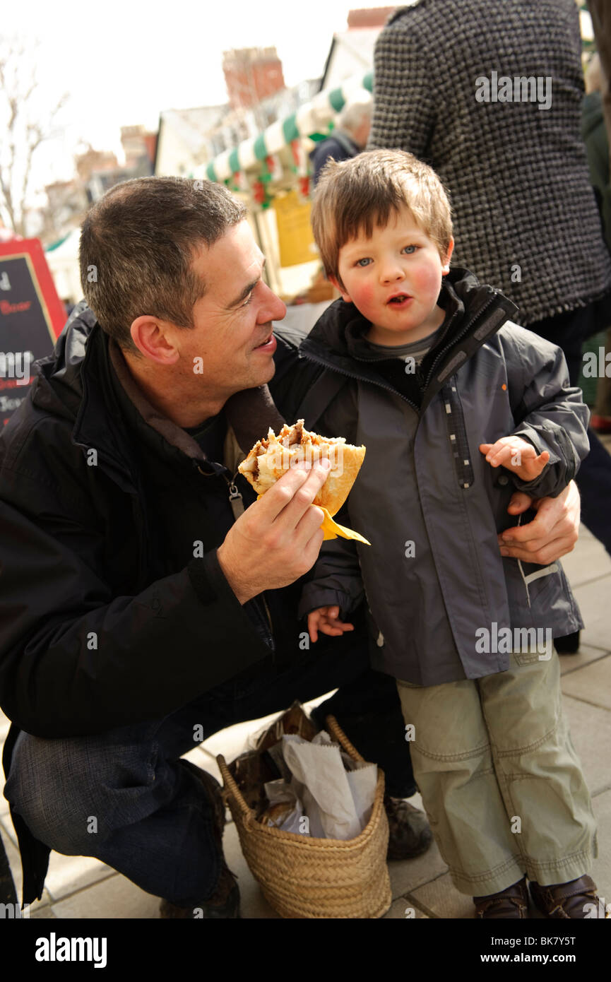 Un padre e figlio giovane mangiare cucinato al momento Organic Wild Boar burger, Aberystwyth Farmers Market, Ceredigion, Wales UK Foto Stock