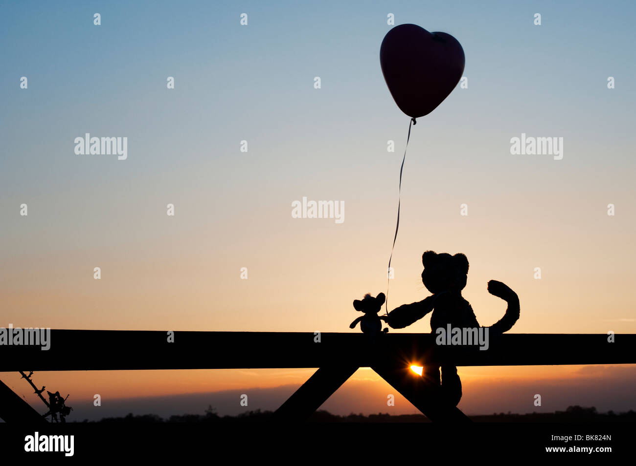 Gatto e topo peluche tenendo a forma di cuore palloncino in amicizia, seduto su un cancello al tramonto. Silhouette Foto Stock