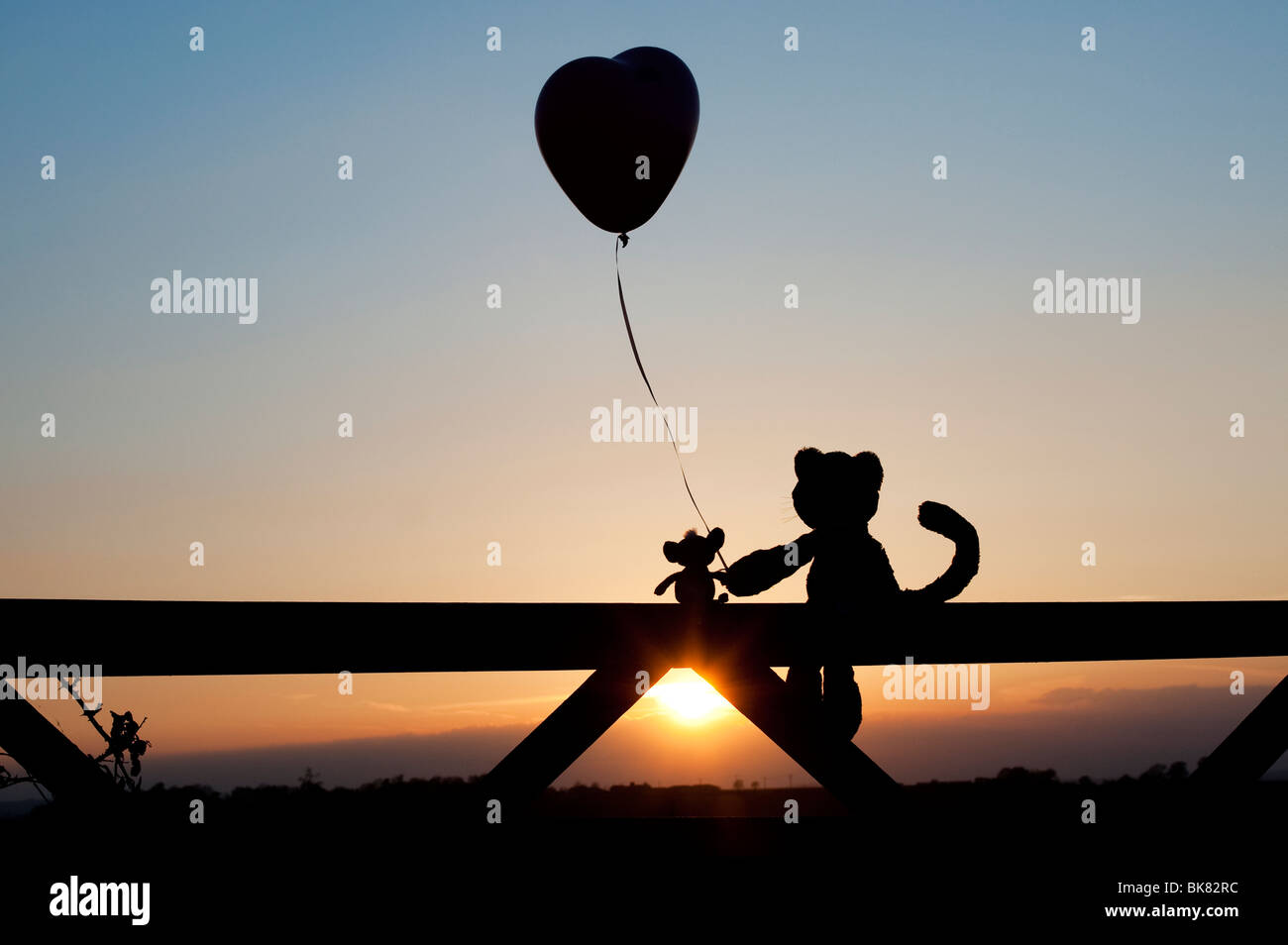 Gatto e topo peluche tenendo a forma di cuore palloncino in amicizia, seduto su un cancello al tramonto. Silhouette Foto Stock