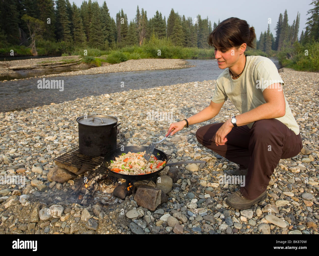 Giovane donna di cottura, friggere il pollo stir fry su un fuoco di campo, ghiaia bar, superiore Liard River, Yukon Territory, Canada Foto Stock