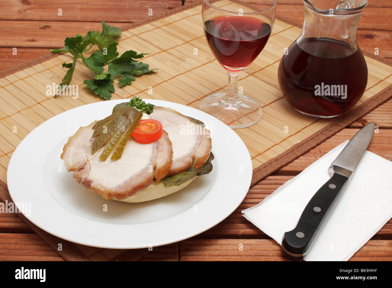 Laminati di arrosto di maiale in sandwich con una ventola cetriolino e fette di pomodoro ciliegino, caraffa e bicchiere di vino rosso, coltello Foto Stock