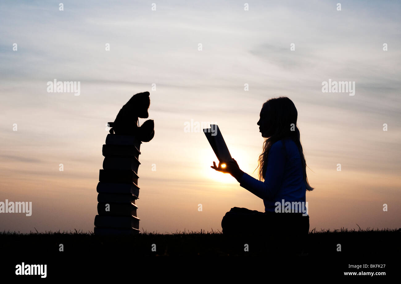 Giovane ragazza di sedersi a leggere un libro di fronte a un orsacchiotto seduto su una pila di libri al tramonto. Silhouette Foto Stock